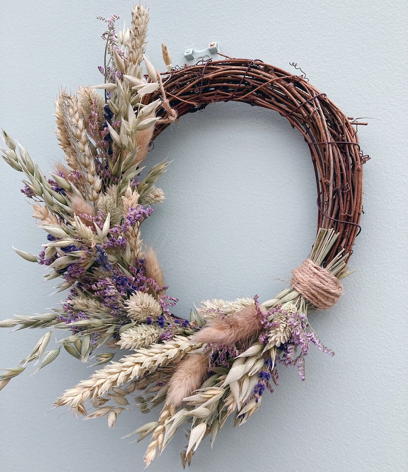 Op de afbeelding: Een krans van gedroogde bloemen met een bruine wijnstokbasis. De krans is versierd met gedroogde lavendel, tarwe en andere grassen.
