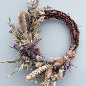 Op de afbeelding: Een krans van gedroogde bloemen met een bruine wijnstokbasis. De krans is versierd met gedroogde tarwe, haver, lavendel en andere gedroogde bloemen in beige, bruin en paarse tinten.