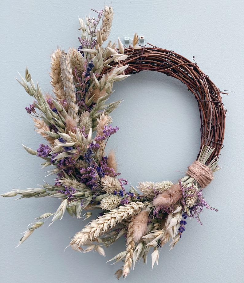 Op de afbeelding: Een krans van gedroogde bloemen met een bruine wijnstokbasis. De krans is versierd met gedroogde tarwe, haver, lavendel en andere gedroogde bloemen in beige, bruin en paarse tinten.