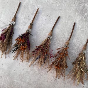 May include: Five small decorative brooms arranged in a row, each featuring a wooden handle, dried flowers in shades of purple, orange, and tan, and a silver-toned pentacle charm tied with twine at the base of the handle.
