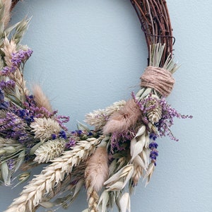 Op de afbeelding: Een krans van gedroogde bloemen met een bruine wijnstokbasis. De krans is versierd met gedroogde tarwe, haver, lavendel en andere gedroogde bloemen.