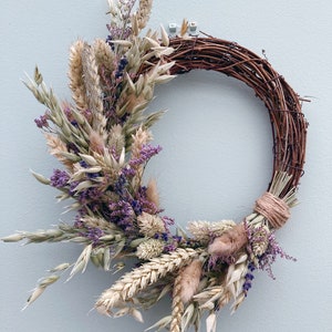 Op de afbeelding: Een krans van gedroogde bloemen met een bruine wijnstokbasis. De krans is versierd met gedroogde tarwe, haver, lavendel en andere gedroogde bloemen in beige, bruin en paarse tinten.