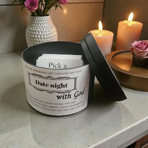 May include: A black and white container with the text "Date night with God" and "Pick a" on the side. The container is open, revealing cards inside. Two lit candles and a vase of flowers are in the background.