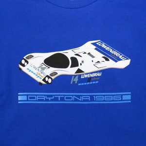 May include: A blue t-shirt with a white and black illustration of a race car. The car has the number 14 and the text "Löwenbräu Spezial" on it. The text "DAYTONA 1986" is printed below the car.