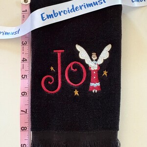 May include: Black hand towel with embroidered red "Joy" text and an angel design. The towel is approximately 9 inches long and has a fringed edge. A white ribbon with the text "Embroiderimust" is draped across the top.