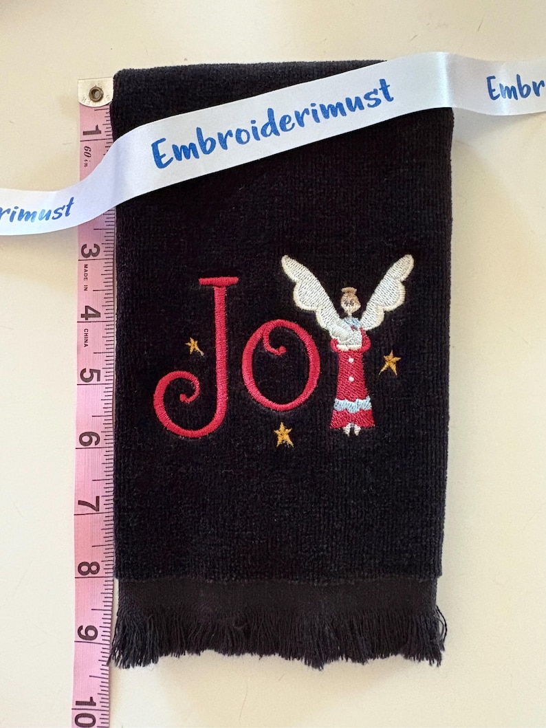 May include: Black hand towel with embroidered red "Joy" text and an angel design. The towel is approximately 9 inches long and has a fringed edge. A white ribbon with the text "Embroiderimust" is draped across the top.