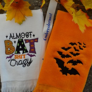 Może przedstawiać: Dwa ręczniki kuchenne z motywami Halloween. Jeden ręcznik biały z tekstem "Almost Bat Shit Crazy" w kolorowym hafcie. Drugi ręcznik jest pomarańczowy z haftowanymi czarnymi nietoperzami.