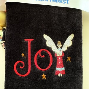 May include: A black towel embroidered with the word "Joy" in red script, accompanied by a small angel figure and gold stars. A white ribbon with the text "Embroiderimust" is draped across the top.