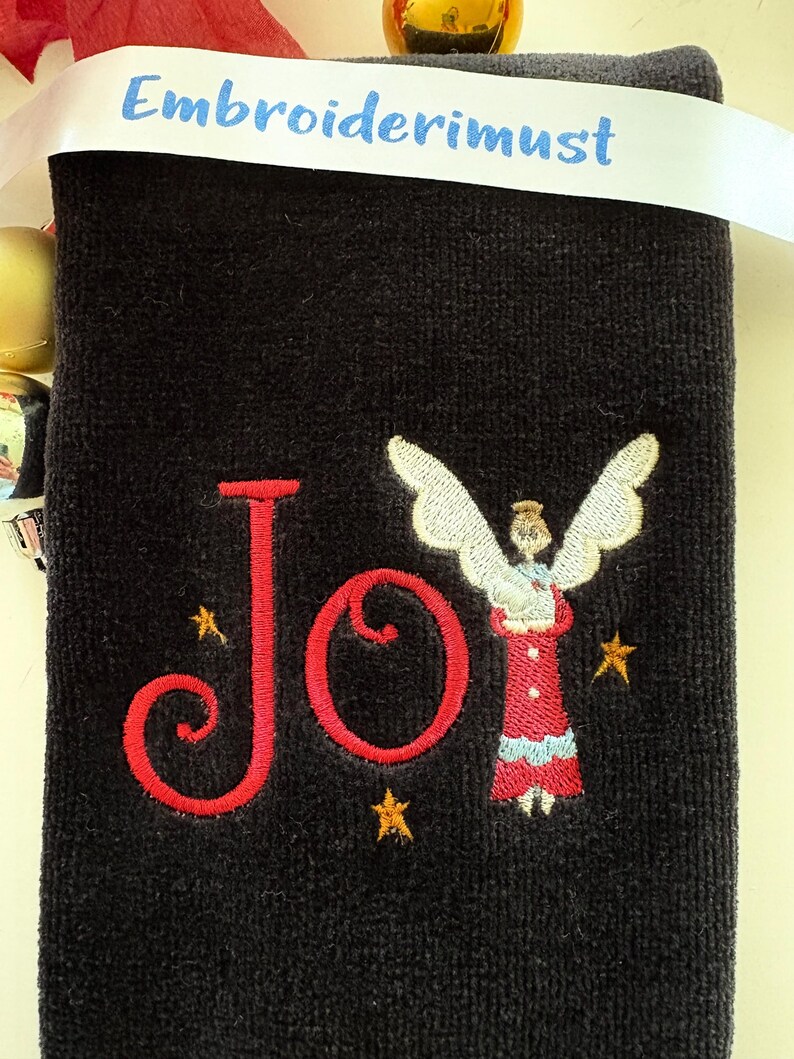 May include: A black towel embroidered with the word "Joy" in red script, accompanied by a small angel figure and gold stars. A white ribbon with the text "Embroiderimust" is draped across the top.