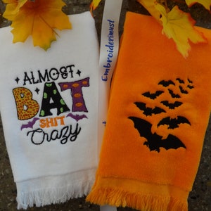 Może przedstawiać: Dwa ręczniki kuchenne w stylu Halloween. Jeden biały ręcznik z napisem "Almost Bat Shit Crazy" w kolorowych haftowanych literach. Drugi ręcznik jest pomarańczowy z czarnym haftowanym wzorem nietoperzy.
