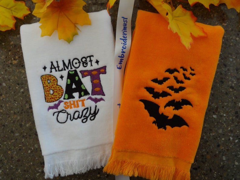Może przedstawiać: Dwa ręczniki kuchenne w stylu Halloween. Jeden biały ręcznik z napisem "Almost Bat Shit Crazy" w kolorowych haftowanych literach. Drugi ręcznik jest pomarańczowy z czarnym haftowanym wzorem nietoperzy.