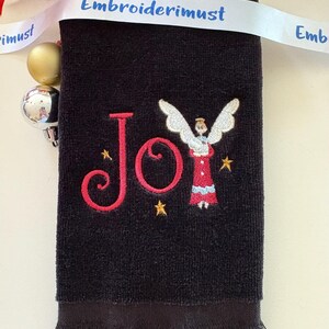 May include: Black hand towel with embroidered red "Joy" text and an angel design. The angel is wearing a red dress and holding a book. Gold stars are embroidered around the angel. A white ribbon with the text "Embroiderimust" is draped across the top.