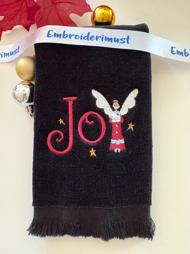 May include: Black hand towel with embroidered red "Joy" text and an angel design. The angel is wearing a red dress and holding a book. Gold stars are embroidered around the angel. A white ribbon with the text "Embroiderimust" is draped across the top.