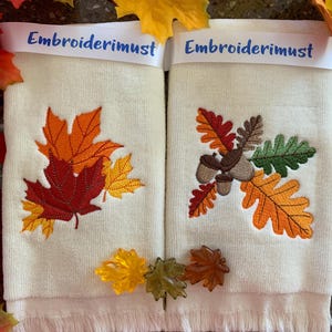 May include: Two white hand towels with embroidered autumn leaves and acorns. The towels have a fringe border and the words "Embroiderimust" are printed on the top of each towel.