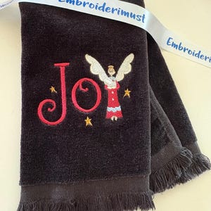 May include: A dark gray towel with embroidered text and imagery. The word "Joy" is embroidered in red, with an angel and gold stars. A white ribbon with the text "Embroiderimust" is draped across the towel.