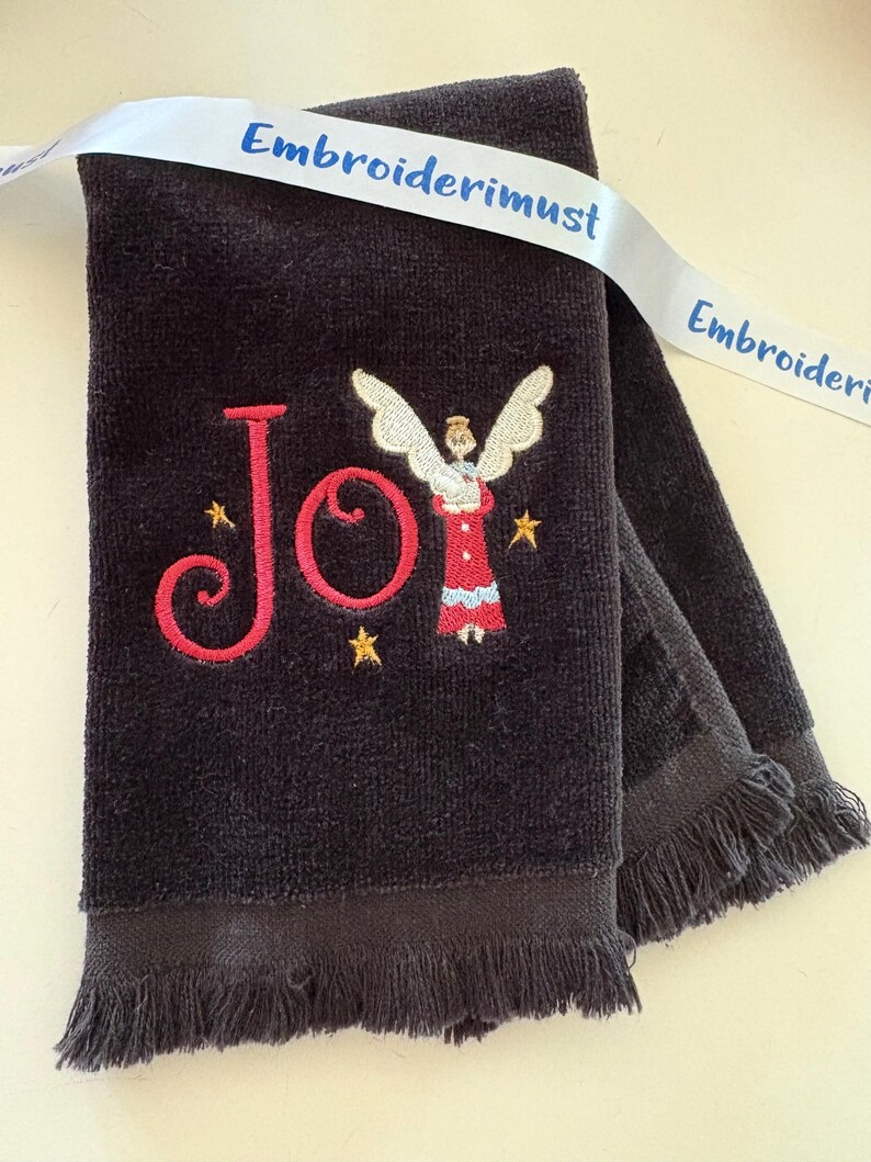 May include: A dark gray towel with embroidered text and imagery. The word "Joy" is embroidered in red, with an angel and gold stars. A white ribbon with the text "Embroiderimust" is draped across the towel.