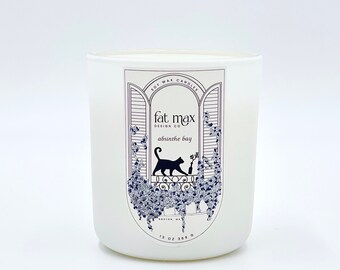 May include: White soy wax candle in a glass jar with a black and white label. The label features a cat sitting on a windowsill with a design of a window with shutters and vines. The label text reads "fat max design co. absinthe bay 13 oz 369 g".
