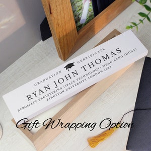 May include: A white plaque with black text reads "Graduation Certificate Ryan John Thomas Aerospace Engineering (Space Technology) MENG/BENG (Hons) Kingston University London 2023". A black graduation cap icon is above the text. The plaque is in a wooden box with a gold tassel.