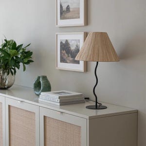 K&ouml;nnte beinhalten: Ein wei&szlig;er Schrank mit zwei T&uuml;ren und geflochtenen Schilfplatten. Der Schrank hat eine hellgraue Arbeitsplatte mit einer schwarzen Tischlampe mit einem geflochtenen Schirm, einer gr&uuml;nen Vase und einem Stapel B&uuml;cher.