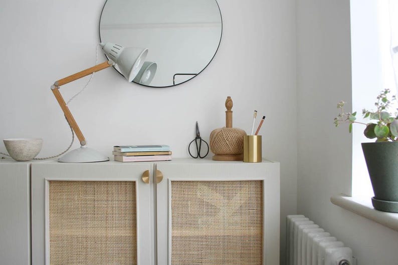 May include: A white cabinet with two doors and a woven cane panel insert. The cabinet has gold hardware and sits on a white countertop. A desk lamp with a white shade and wooden arm is on the countertop. Other items on the countertop include a stack of books, a spool of twine, a gold pencil holder, and a pair of scissors.