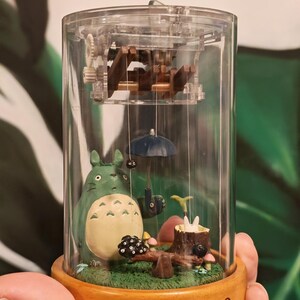 May include: A clear, cylindrical music box featuring a scene from the animated film "My Neighbor Totoro." The scene includes a green Totoro figure, a blue umbrella, and a wooden base with the word "TOTORO" printed on it.
