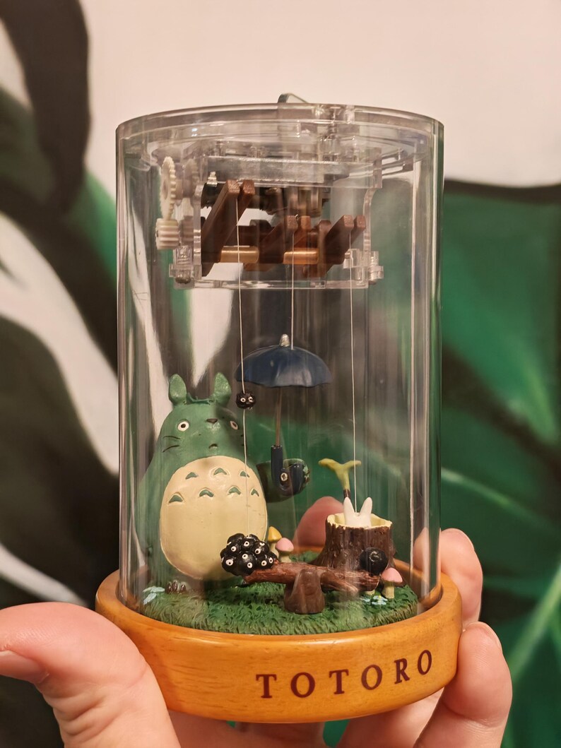 May include: A clear, cylindrical music box featuring a scene from the animated film "My Neighbor Totoro." The scene includes a green Totoro figure, a blue umbrella, and a wooden base with the word "TOTORO" printed on it.