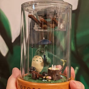 May include: A clear, cylindrical music box featuring a green Totoro figure holding a blue umbrella. The base is wooden with the word "TOTORO" in brown. Inside, a miniature scene includes a tree stump and mushrooms.