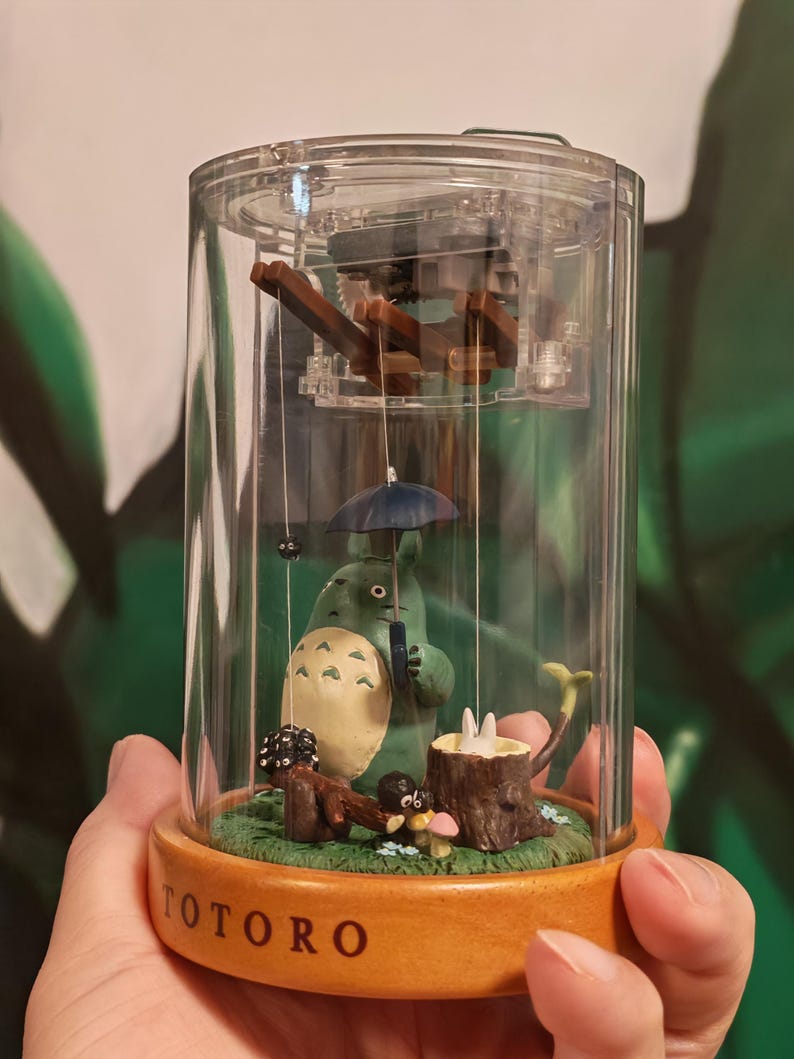 May include: A clear, cylindrical music box featuring a green Totoro figure holding a blue umbrella. The base is wooden with the word "TOTORO" in brown. Inside, a miniature scene includes a tree stump and mushrooms.