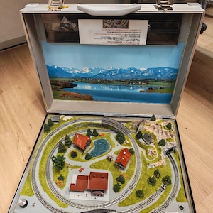 Puede incluir: Un tren en miniatura dentro de un maletín plateado. El conjunto presenta una vía circular, pequeños edificios con tejados rojos, árboles y un estanque. El fondo muestra un paisaje escénico con montañas y un lago.