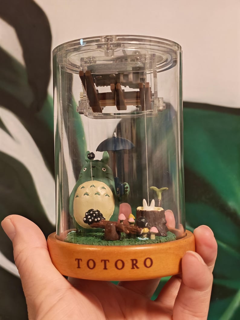 May include: A Totoro-themed decorative snow globe. The clear, cylindrical globe houses a miniature scene with the character Totoro, mushrooms, and a small umbrella. The base is wooden and has the word "TOTORO" printed on it.