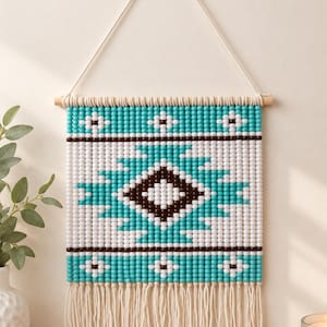 May include: A rectangular wall hanging made of small turquoise, white, and dark brown beads arranged in a geometric diamond pattern. The piece is suspended from a wooden dowel by a white cord and features a fringe of white cotton tassels at the bottom.
