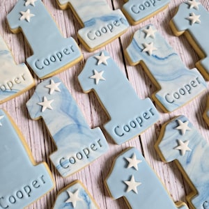 May include: A collection of number one cookies, decorated with light blue icing and white star accents. Each cookie has the name "Cooper" imprinted on it. Some cookies have a marbled blue and white design.