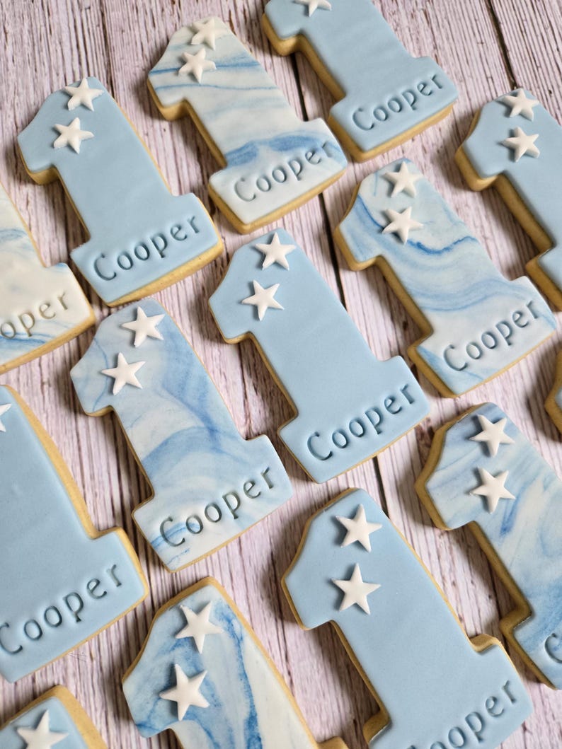 May include: A collection of number one cookies, decorated with light blue icing and white star accents. Each cookie has the name "Cooper" imprinted on it. Some cookies have a marbled blue and white design.