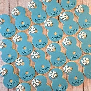 May include: A collection of number eight-shaped cookies with light blue frosting. Each cookie has the name "Amara" written on it and is decorated with a white snowflake. The cookies are arranged on a light wooden surface.