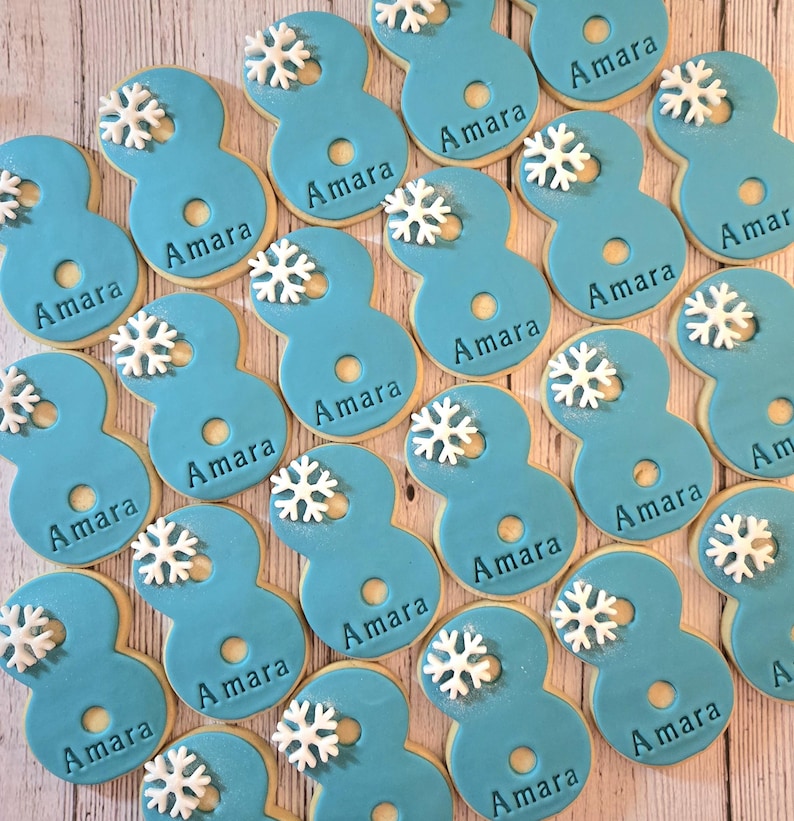 May include: A collection of number eight-shaped cookies with light blue frosting. Each cookie has the name "Amara" written on it and is decorated with a white snowflake. The cookies are arranged on a light wooden surface.