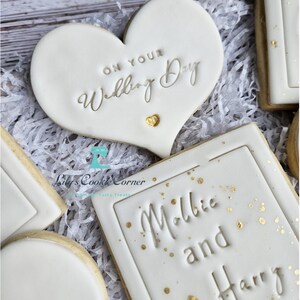 Pode incluir: Biscoitos brancos com glac&ecirc; e detalhes dourados. Um biscoito em forma de cora&ccedil;&atilde;o com o texto "ON YOUR Wedding Day". Outro biscoito retangular com os nomes "Millie and Harry". Os biscoitos est&atilde;o em papel branco.