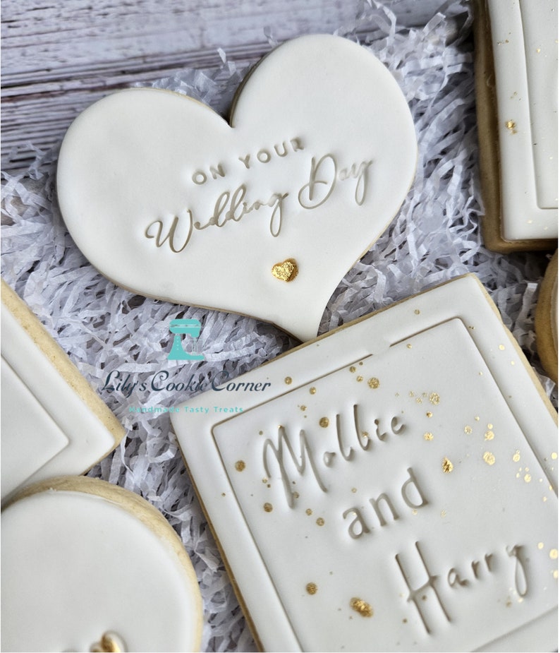 Pode incluir: Biscoitos brancos com glac&ecirc; e detalhes dourados. Um biscoito em forma de cora&ccedil;&atilde;o com o texto "ON YOUR Wedding Day". Outro biscoito retangular com os nomes "Millie and Harry". Os biscoitos est&atilde;o em papel branco.