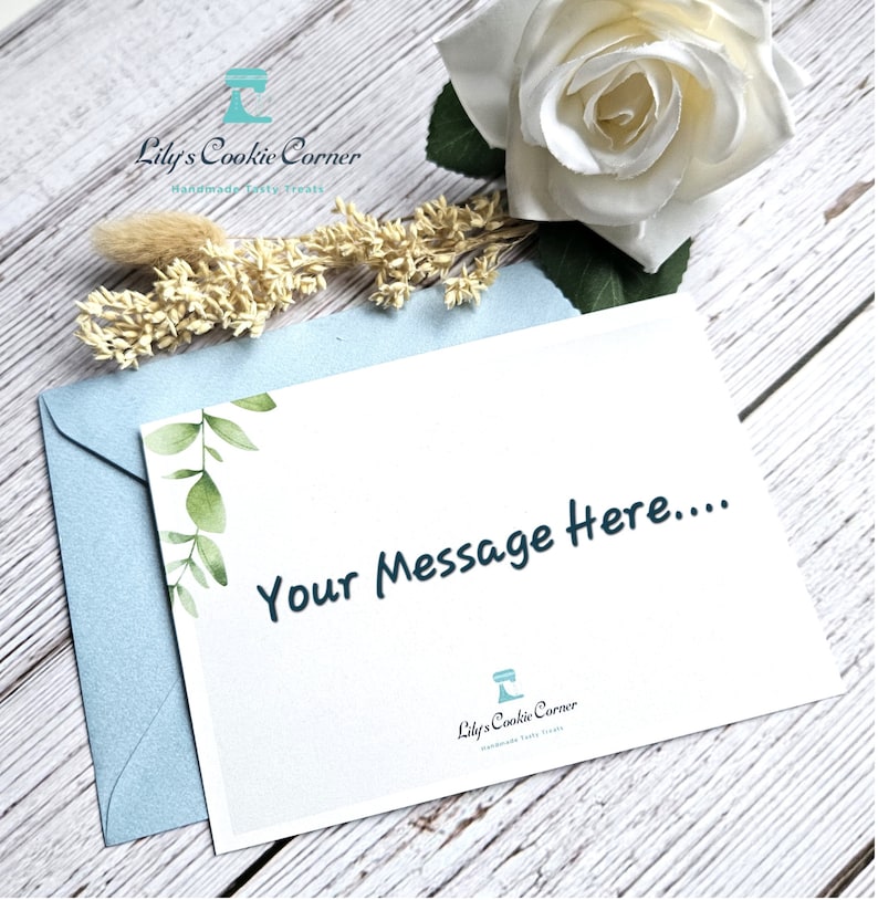 Pode incluir: Um cart&atilde;o de felicita&ccedil;&otilde;es branco com o texto "Your Message Here...." &eacute; colocado em cima de um envelope azul claro. O cart&atilde;o tem um desenho de folhas verdes no canto. Uma rosa branca e flores secas no fundo. O logotipo da Lily's Cookie Corner est&aacute; em baixo.