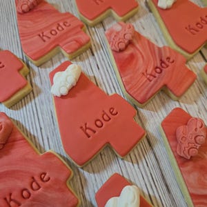 May include: Red frosted cookies with the name "Kode" imprinted on them. The cookies have a marbled design and are decorated with white icing accents. The cookies are arranged on a wooden surface.