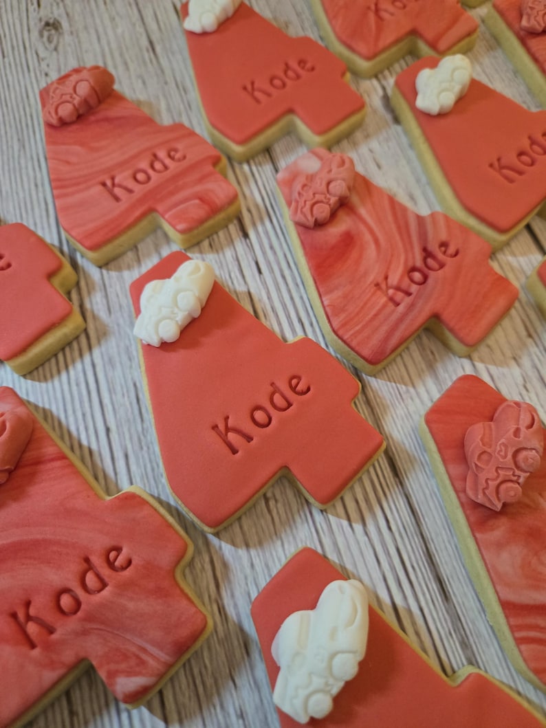 May include: Red frosted cookies with the name "Kode" imprinted on them. The cookies have a marbled design and are decorated with white icing accents. The cookies are arranged on a wooden surface.