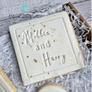 Pode incluir: Um biscoito quadrado com glac&ecirc; branco, com os nomes "Millie and Harry" em relevo e decorado com salpicos dourados. O biscoito &eacute; apresentado numa caixa castanha com papel picado. O log&oacute;tipo "Lily's Cookie Corner" &eacute; vis&iacute;vel.