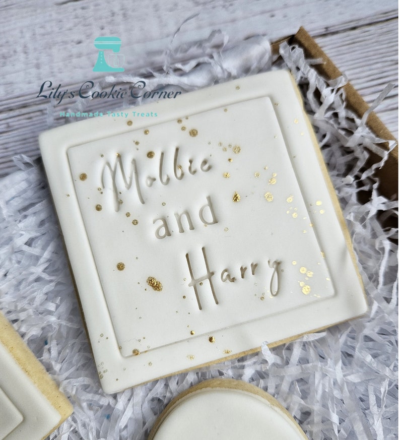 Pode incluir: Um biscoito quadrado com glac&ecirc; branco, com os nomes "Millie and Harry" em relevo e decorado com salpicos dourados. O biscoito &eacute; apresentado numa caixa castanha com papel picado. O log&oacute;tipo "Lily's Cookie Corner" &eacute; vis&iacute;vel.