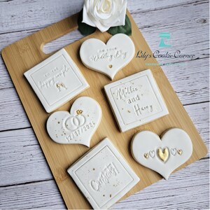 Pode incluir: Uma cole&ccedil;&atilde;o de biscoitos brancos decorados, dispostos numa t&aacute;bua de madeira. Os biscoitos, em forma de cora&ccedil;&atilde;o e quadrados, apresentam detalhes dourados e inscri&ccedil;&otilde;es como "To the happy couple" e "On your Wedding Day".
