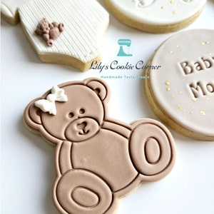 May include: A set of four decorated sugar cookies. One cookie is shaped like a baby onesie with a small teddy bear on the front. Another cookie is round with the words "Oh Baby" in gold lettering. A third cookie is round with the words "Baby Moon" in gold lettering. The fourth cookie is shaped like a teddy bear with a white bow.