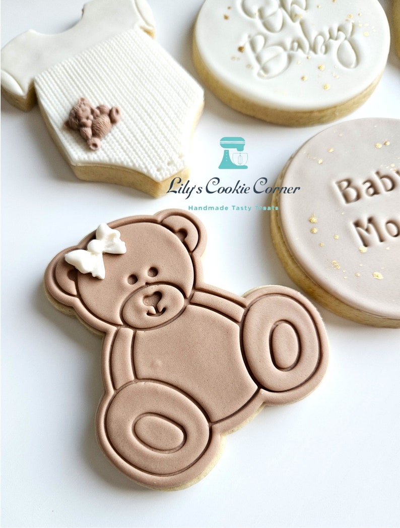 May include: A set of four decorated sugar cookies. One cookie is shaped like a baby onesie with a small teddy bear on the front. Another cookie is round with the words "Oh Baby" in gold lettering. A third cookie is round with the words "Baby Moon" in gold lettering. The fourth cookie is shaped like a teddy bear with a white bow.