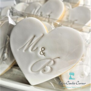 May include: Heart-shaped cookies with white icing and the letters "M & B" embossed on the surface. The cookies have a yellow edge and are packaged in clear cellophane. The image also includes the text "Lily's Cookie Corner" and "Handmade Tasty Treats".