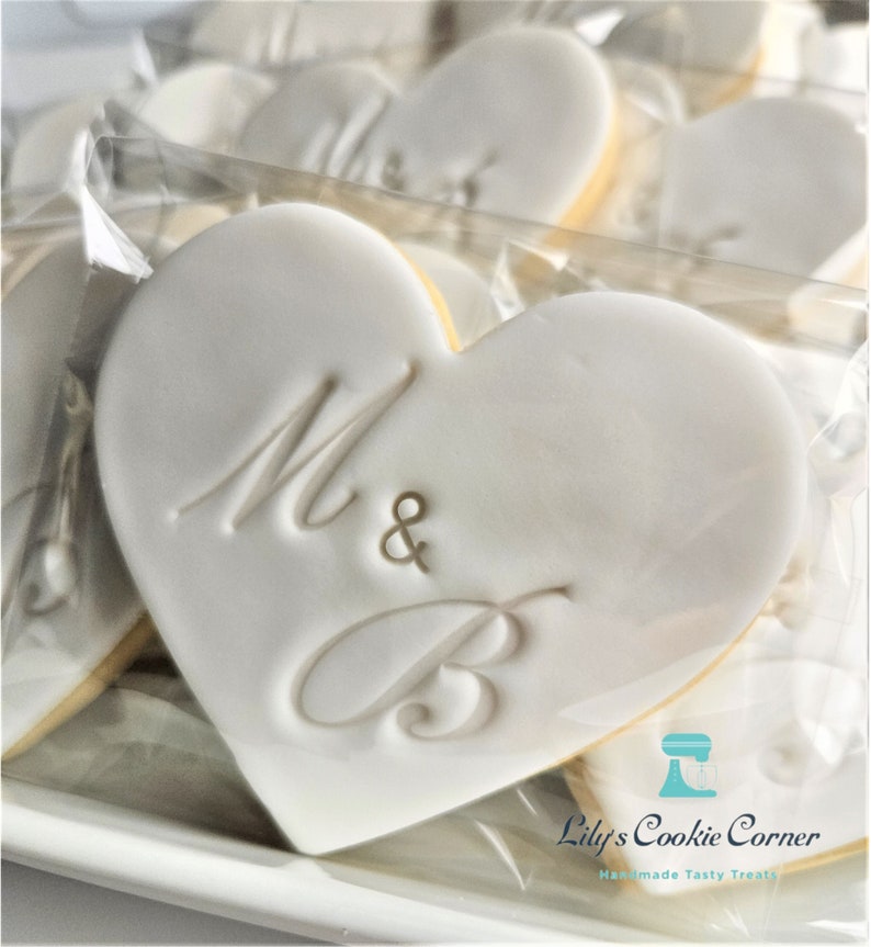 May include: Heart-shaped cookies with white icing and the letters "M & B" embossed on the surface. The cookies have a yellow edge and are packaged in clear cellophane. The image also includes the text "Lily's Cookie Corner" and "Handmade Tasty Treats".