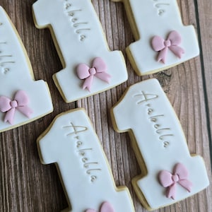 May include: White frosted number one cookies with the name "Arabella" written on them. Each cookie has a pink bow at the bottom. The cookies are on a wooden surface.