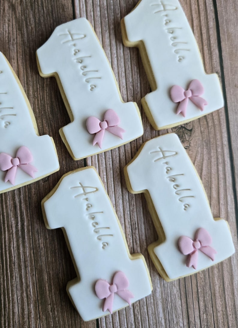 May include: White frosted number one cookies with the name "Arabella" written on them. Each cookie has a pink bow at the bottom. The cookies are on a wooden surface.