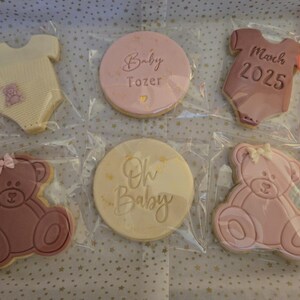 May include: Six decorated sugar cookies in a gift box. The cookies are pink and white and feature a baby onesie, a teddy bear, and the text "Baby Tozer", "Oh Baby", and "March 2025".